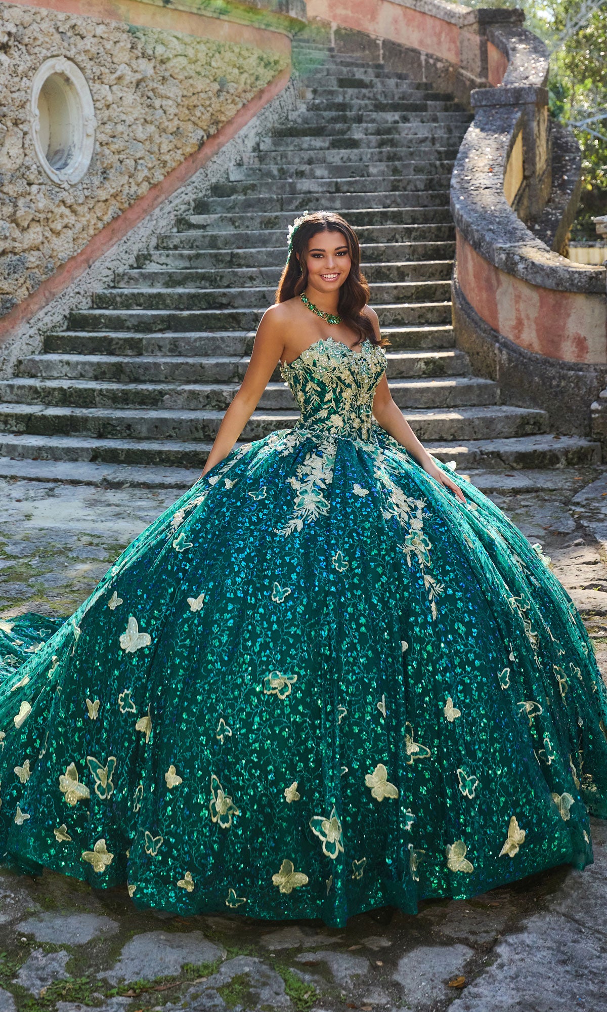 A woman stands outdoors on stone steps, wearing the Strapless Quinceanera Dress: Princesa PR30121, an elaborate green gown with gold embroidered lace and butterfly details.